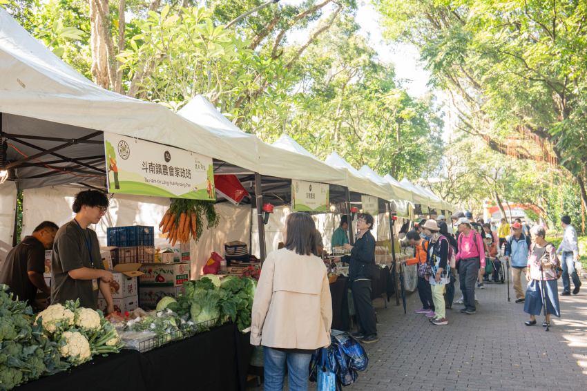 臺大USR農夫市集現場人潮踴躍,民眾在攤位前選購新鮮蔬菜,攤位上掛著紅蘿蔔,展現農村活力。